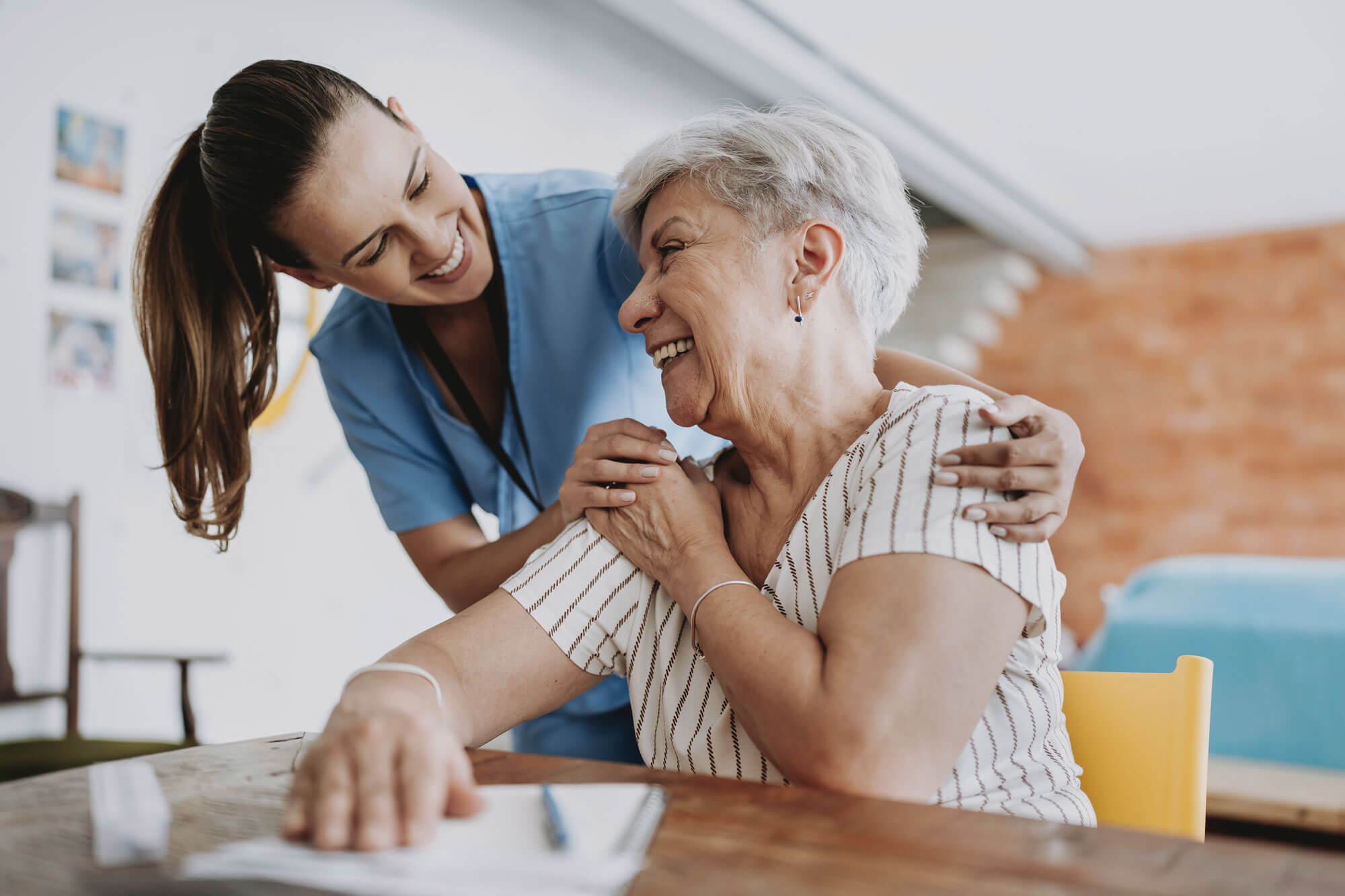 A young caregiver in blue scrubs smiles and gently hugs an elderly woman with short gray hair. They are seated at a table, sharing a joyful moment. The elderly woman is looking up at the caregiver, holding her hand.