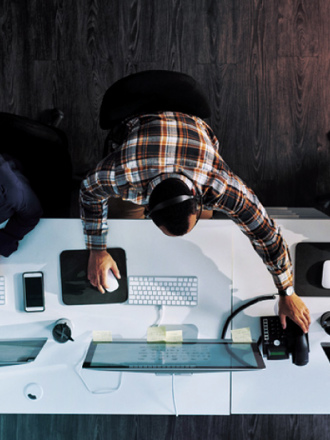 Overhead view of three office workers seated at a long desk with computers. One wears headphones and uses a mouse, the others type on keyboards. The desk holds phones, monitors, and personal items. Dark wood flooring is visible.