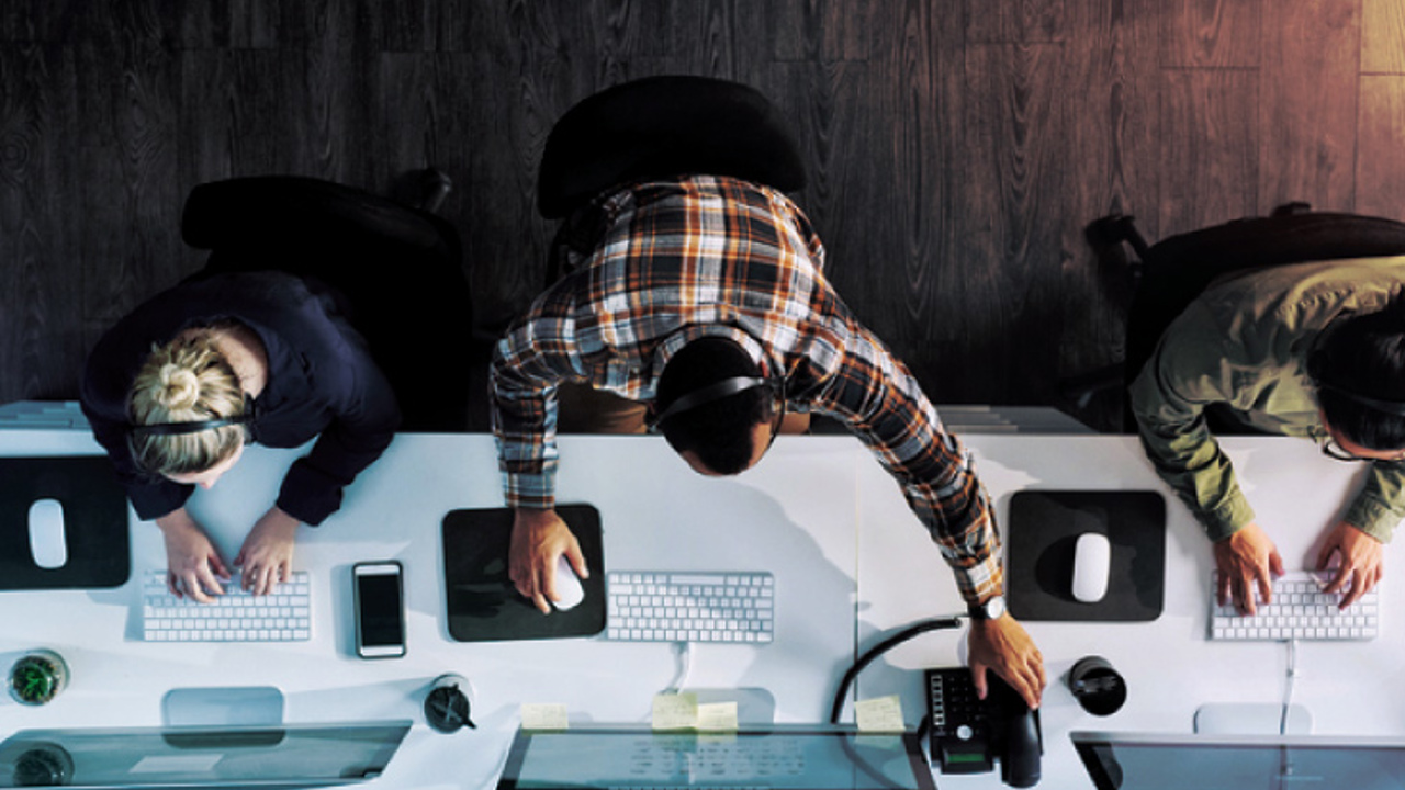 Overhead view of three office workers seated at a long desk with computers. One wears headphones and uses a mouse, the others type on keyboards. The desk holds phones, monitors, and personal items. Dark wood flooring is visible.