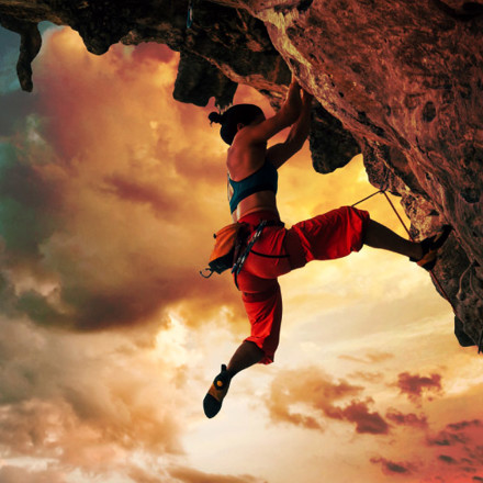 Athletic woman climbing on overhanging cliff rock with sunset sky background.