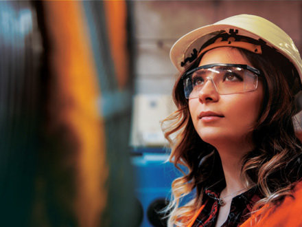 A woman wearing a white hard hat and safety glasses looks upward with focus. She is in an industrial setting, wearing an orange safety vest. The background is blurred.