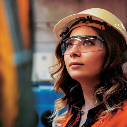 A woman wearing a white hard hat and safety glasses looks upward with focus. She is in an industrial setting, wearing an orange safety vest. The background is blurred.