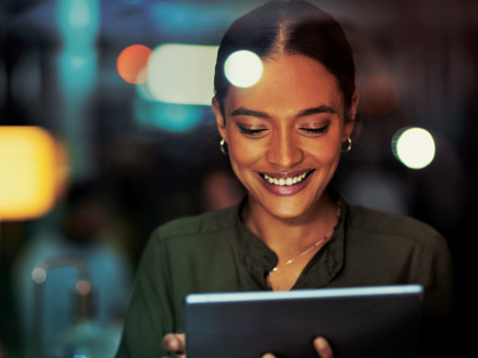 A woman is smiling while looking at a tablet in a dimly lit environment. She is wearing a dark green blouse and hoop earrings. The background is blurred with soft lights.