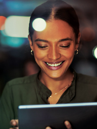 A woman is smiling while looking at a tablet in a dimly lit environment. She is wearing a dark green blouse and hoop earrings. The background is blurred with soft lights.