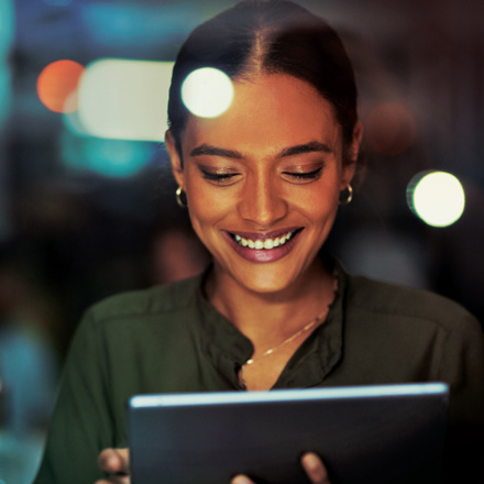 A woman is smiling while looking at a tablet in a dimly lit environment. She is wearing a dark green blouse and hoop earrings. The background is blurred with soft lights.