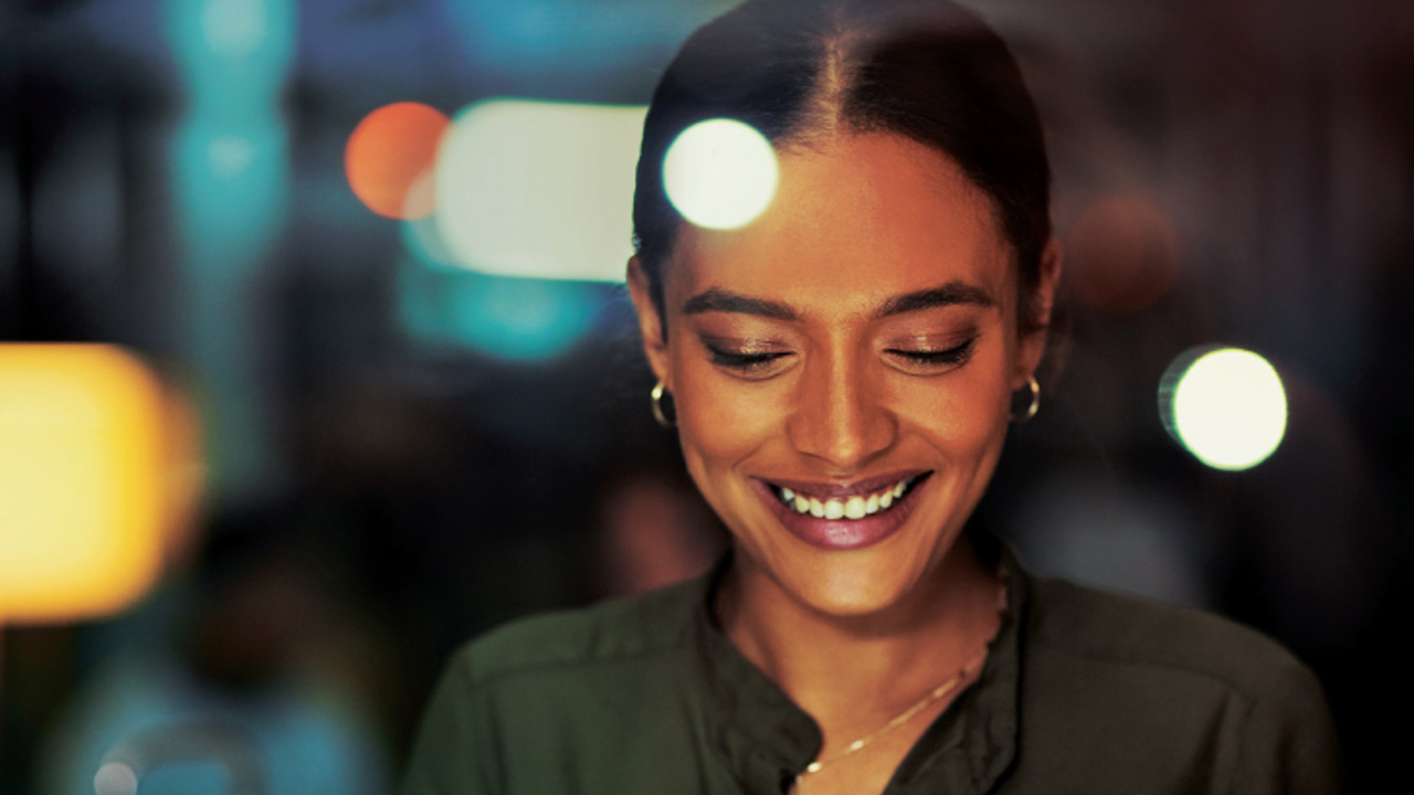 A woman is smiling while looking at a tablet in a dimly lit environment. She is wearing a dark green blouse and hoop earrings. The background is blurred with soft lights.