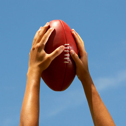 Hands holding Australian football up to the sky