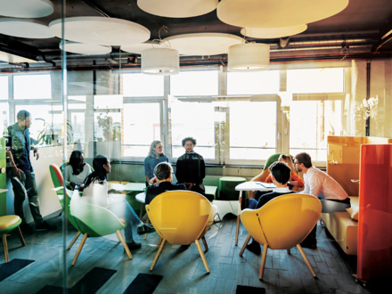 A modern office lounge with people engaged in a meeting. The room features bright, colorful chairs, round ceiling lights, and large windows allowing natural light. Participants are seated around a table, and a few are standing in conversation.