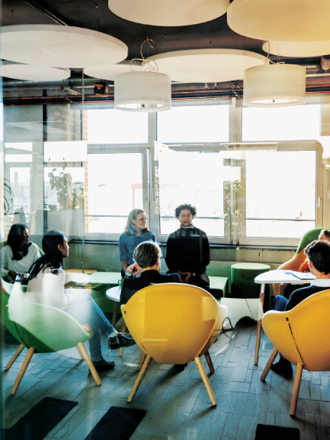 A modern office lounge with people engaged in a meeting. The room features bright, colorful chairs, round ceiling lights, and large windows allowing natural light. Participants are seated around a table, and a few are standing in conversation.