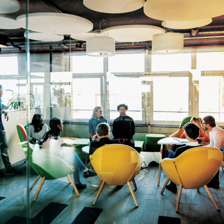 A modern office lounge with people engaged in a meeting. The room features bright, colorful chairs, round ceiling lights, and large windows allowing natural light. Participants are seated around a table, and a few are standing in conversation.