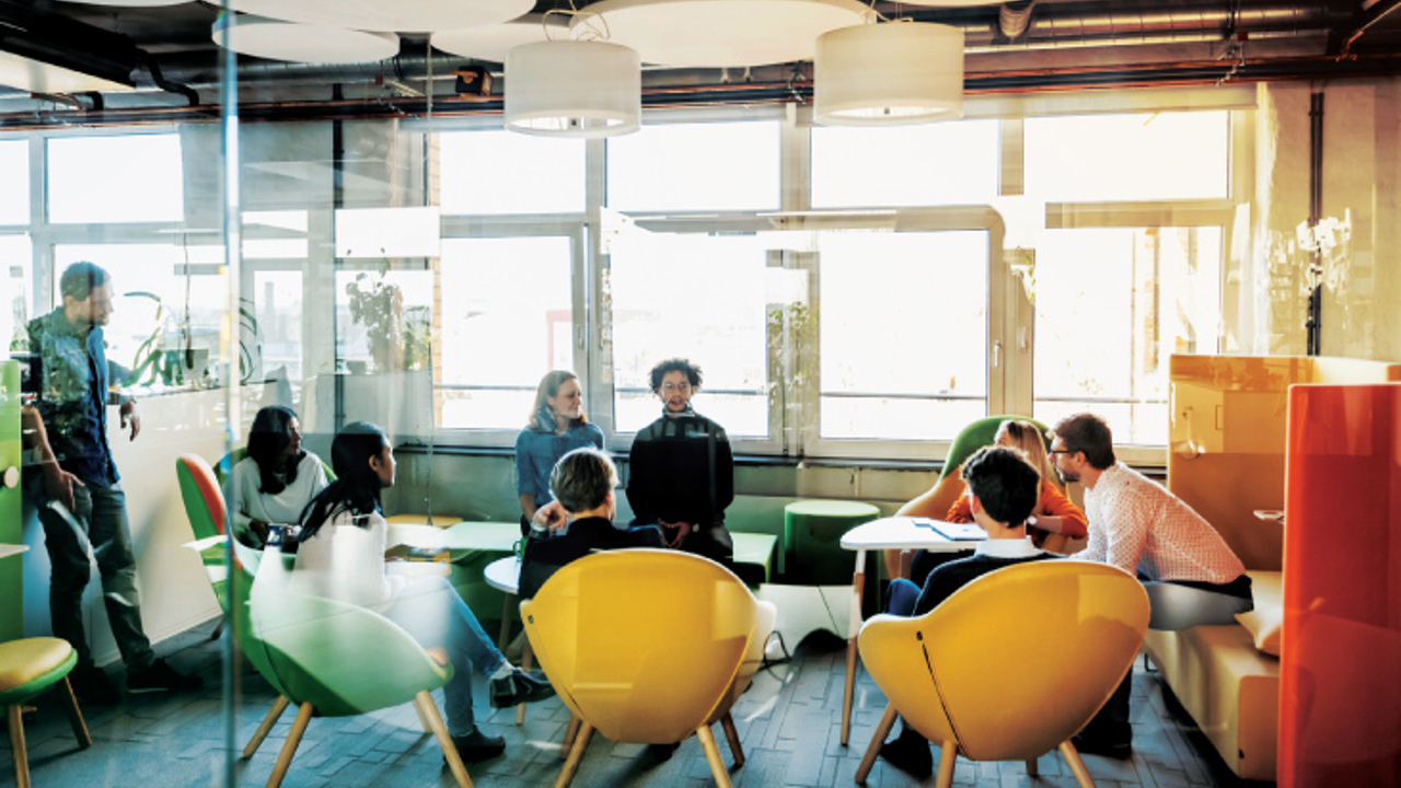 A modern office lounge with people engaged in a meeting. The room features bright, colorful chairs, round ceiling lights, and large windows allowing natural light. Participants are seated around a table, and a few are standing in conversation.