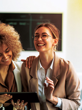 Two women are smiling while looking at a tablet. One has curly hair and is wearing a white blazer, and the other has glasses, a bun, and a tan blazer. They are in a bright room with a blurred chalkboard in the background.