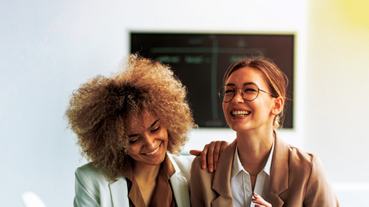 Two women are smiling while looking at a tablet. One has curly hair and is wearing a white blazer, and the other has glasses, a bun, and a tan blazer. They are in a bright room with a blurred chalkboard in the background.