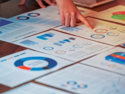 A person points to a document on a table filled with charts and graphs, including bar and pie charts. The papers appear to show financial or business data, with various colors used in the visualizations.