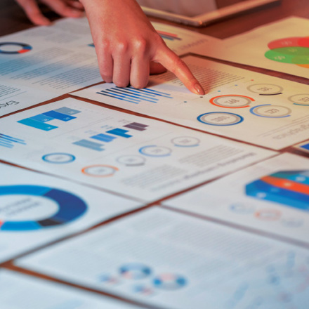 A person points to a document on a table filled with charts and graphs, including bar and pie charts. The papers appear to show financial or business data, with various colors used in the visualizations.