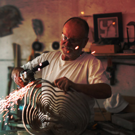 a Senior man using an angle grinder to create sculptures out of metal in his art studio.