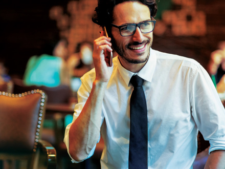 A man in glasses and a white shirt with a black tie is sitting and smiling while talking on the phone in a warmly lit, blurred background setting.