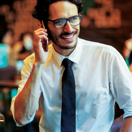 A man in glasses and a white shirt with a black tie is sitting and smiling while talking on the phone in a warmly lit, blurred background setting.