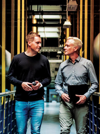 Two men walking through an industrial setting with yellow beams and large ducts. One wears a black sweater and jeans, holding an item, and the other wears a gray shirt with khaki pants, carrying a black folder. They are in conversation.