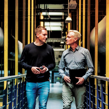 Two men walking through an industrial setting with yellow beams and large ducts. One wears a black sweater and jeans, holding an item, and the other wears a gray shirt with khaki pants, carrying a black folder. They are in conversation.