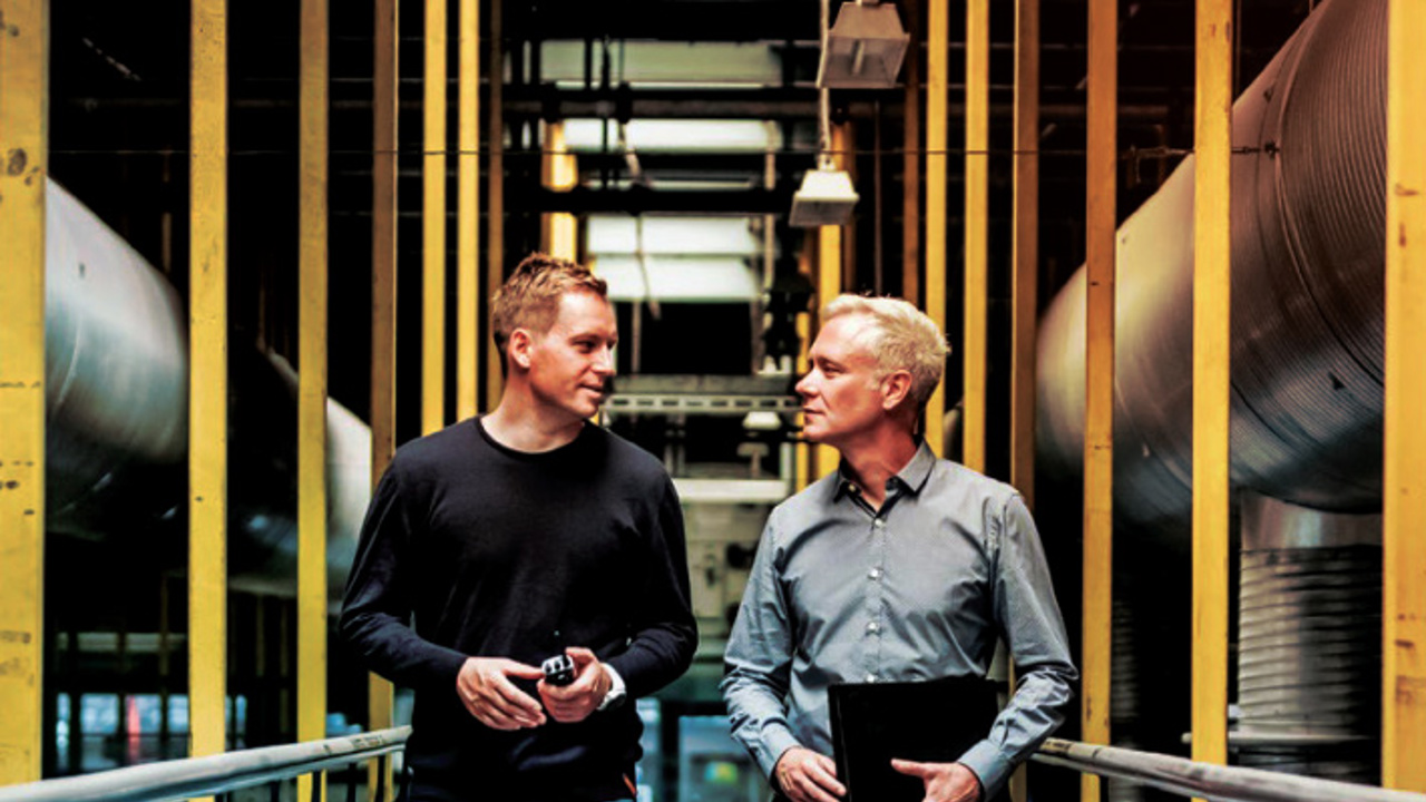 Two men walking through an industrial setting with yellow beams and large ducts. One wears a black sweater and jeans, holding an item, and the other wears a gray shirt with khaki pants, carrying a black folder. They are in conversation.