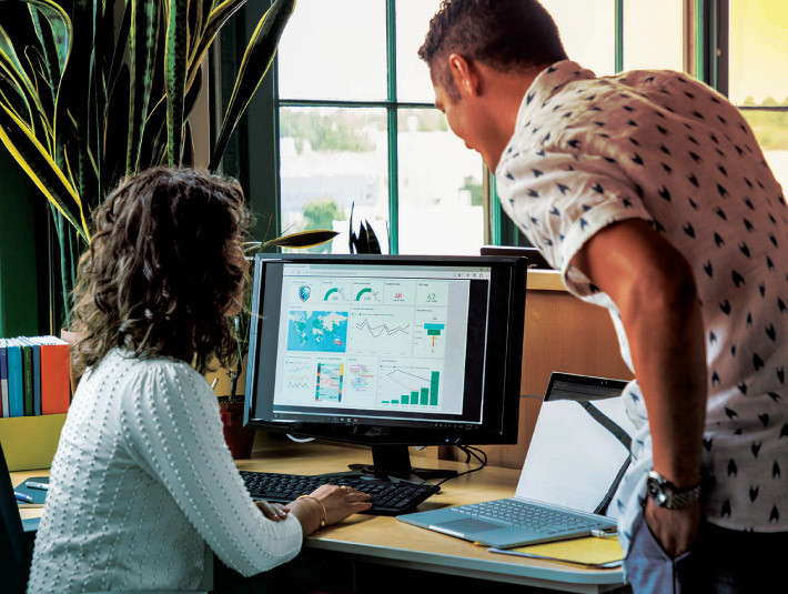 Two people in an office are examining a computer screen displaying various colorful charts and graphs. One person is seated, and the other is standing, leaning in to look closely at the screen. Nearby are a laptop and office supplies.