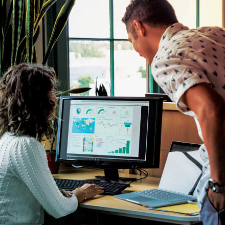 Two people in an office are examining a computer screen displaying various colorful charts and graphs. One person is seated, and the other is standing, leaning in to look closely at the screen. Nearby are a laptop and office supplies.