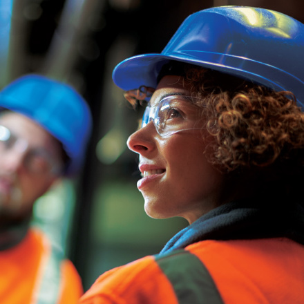 Two co-working in the boiler room, wearing high viz jackets. Female in foreground, looking over her shoulder.