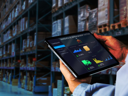 A person is holding a tablet displaying a warehouse management system dashboard with graphs and statistics. The background shows shelves filled with boxes in a warehouse.