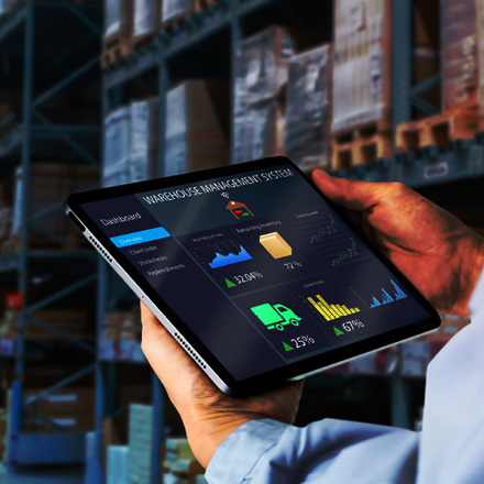 A person is holding a tablet displaying a warehouse management system dashboard with graphs and statistics. The background shows shelves filled with boxes in a warehouse.