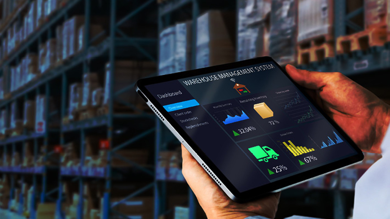 A person is holding a tablet displaying a warehouse management system dashboard with graphs and statistics. The background shows shelves filled with boxes in a warehouse.