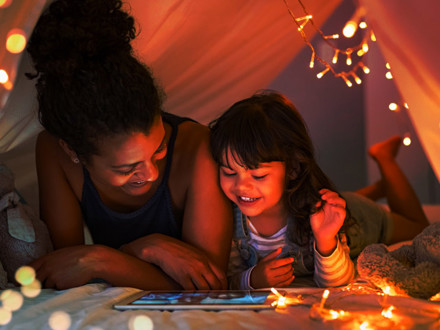 African mother and cute smiling girl using digital tablet while lying in illuminted tent in kid bedroom. Cheerful ethnic woman and lovely daughter on video call under a cozy hut. Lovely little girl with mom watching cartoon on digital tablet in bedroom.