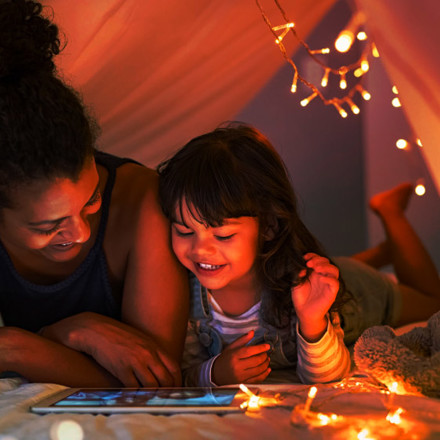 African mother and cute smiling girl using digital tablet while lying in illuminted tent in kid bedroom. Cheerful ethnic woman and lovely daughter on video call under a cozy hut. Lovely little girl with mom watching cartoon on digital tablet in bedroom.