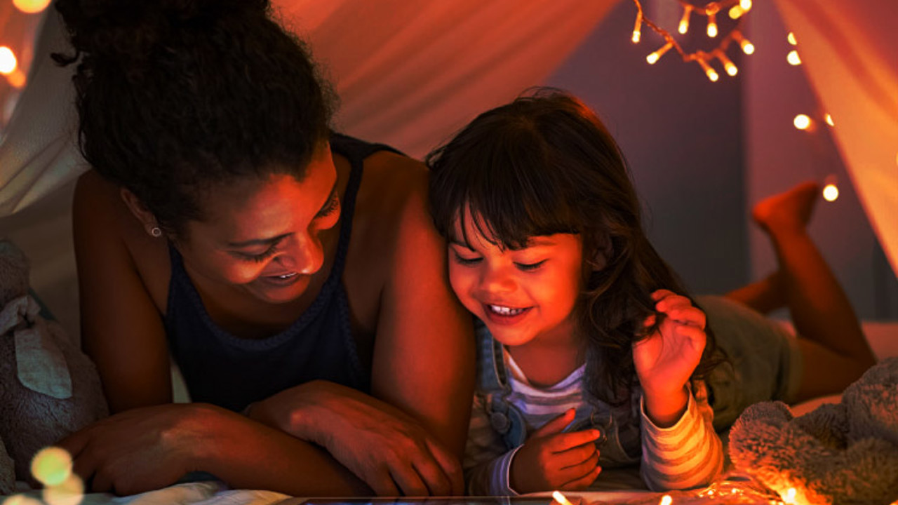 African mother and cute smiling girl using digital tablet while lying in illuminted tent in kid bedroom. Cheerful ethnic woman and lovely daughter on video call under a cozy hut. Lovely little girl with mom watching cartoon on digital tablet in bedroom.