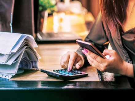 A person using a smartphone and calculator at a desk, surrounded by a stack of papers. The scene suggests a focus on financial tasks or budgeting.