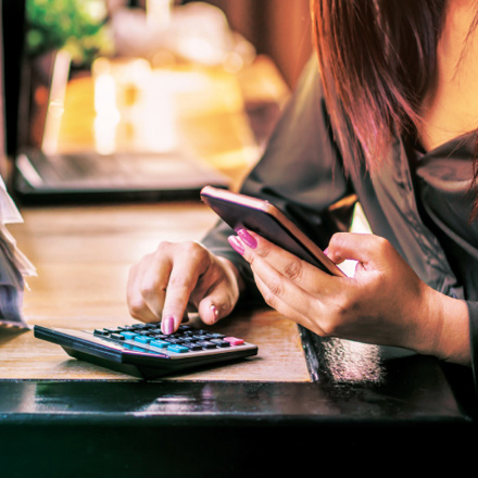 A person using a smartphone and calculator at a desk, surrounded by a stack of papers. The scene suggests a focus on financial tasks or budgeting.