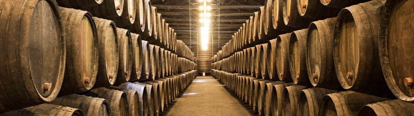 A dimly lit warehouse with wooden barrels stacked in neat rows on either side of a central aisle. The barrels rest on wooden supports, indicating storage of liquids such as wine or spirits. The ceiling features exposed beams and overhead lighting.