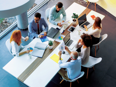 A diverse group of six people sit around a modern conference table with laptops, papers, and coffee cups. They are engaged in discussion, viewing charts and graphs. The setting is bright with natural light and plants on the table.