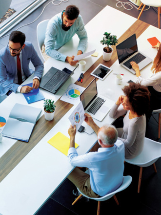 A diverse group of six people sit around a modern conference table with laptops, papers, and coffee cups. They are engaged in discussion, viewing charts and graphs. The setting is bright with natural light and plants on the table.