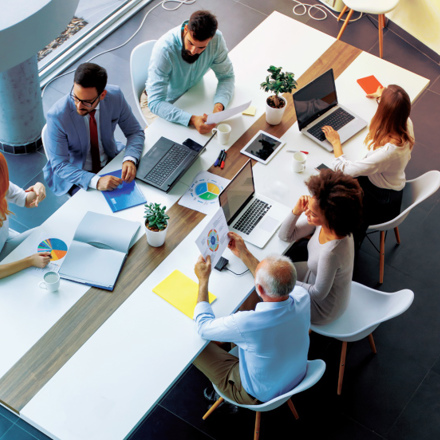 A diverse group of six people sit around a modern conference table with laptops, papers, and coffee cups. They are engaged in discussion, viewing charts and graphs. The setting is bright with natural light and plants on the table.