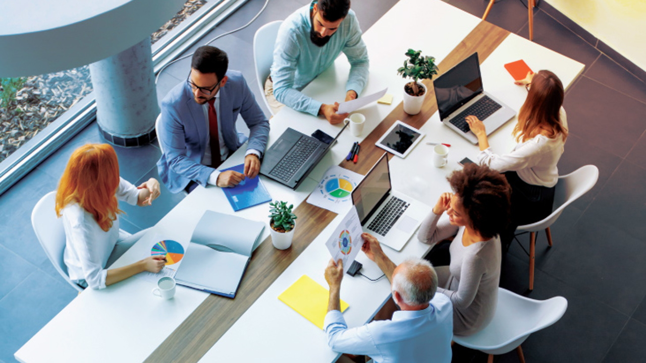 A diverse group of six people sit around a modern conference table with laptops, papers, and coffee cups. They are engaged in discussion, viewing charts and graphs. The setting is bright with natural light and plants on the table.