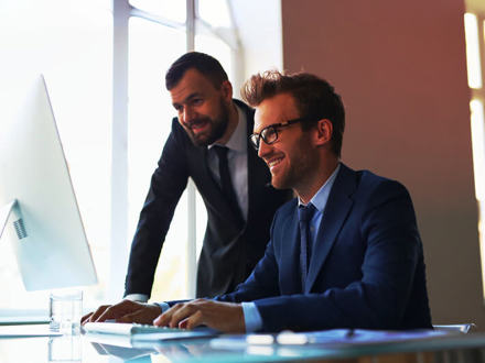 Two men in suits sit at a desk in a brightly lit office. One man with glasses is smiling and typing on a laptop, while the other looks on attentively. A large window in the background lets in natural light.