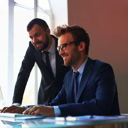 Two men in suits sit at a desk in a brightly lit office. One man with glasses is smiling and typing on a laptop, while the other looks on attentively. A large window in the background lets in natural light.