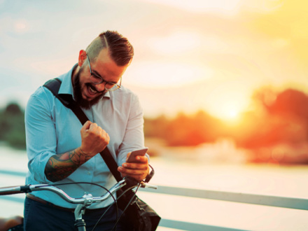 A bearded man with glasses, wearing a light blue shirt, is enthusiastically looking at his smartphone while seated on a bicycle. He clenches his fist in excitement. The background features a sunset over a body of water.