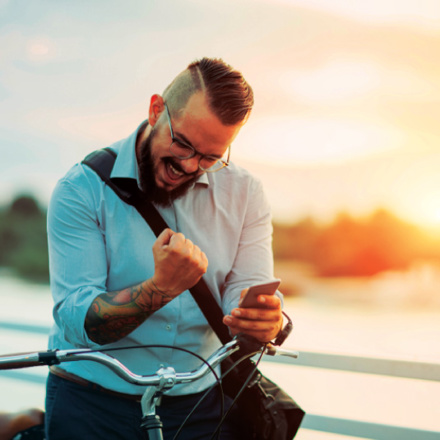 A bearded man with glasses, wearing a light blue shirt, is enthusiastically looking at his smartphone while seated on a bicycle. He clenches his fist in excitement. The background features a sunset over a body of water.