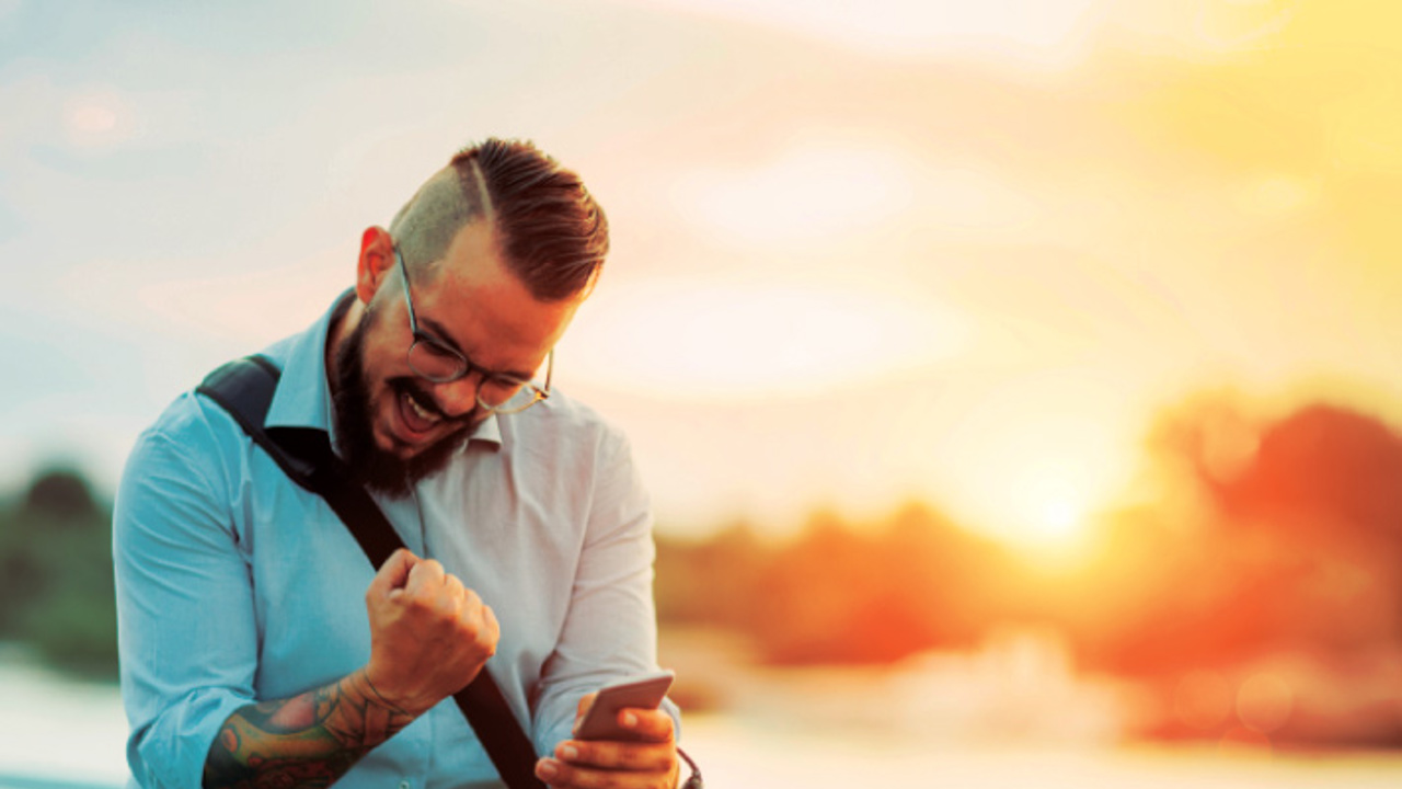 A bearded man with glasses, wearing a light blue shirt, is enthusiastically looking at his smartphone while seated on a bicycle. He clenches his fist in excitement. The background features a sunset over a body of water.