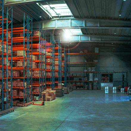 A spacious warehouse with high ceilings, featuring shelves stacked with boxes on the left and open floor space on the right. Sunlight streams in through a skylight, illuminating the scene.