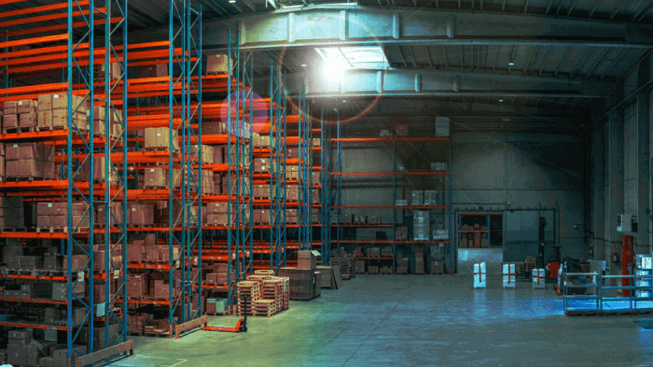 A spacious warehouse with high ceilings, featuring shelves stacked with boxes on the left and open floor space on the right. Sunlight streams in through a skylight, illuminating the scene.