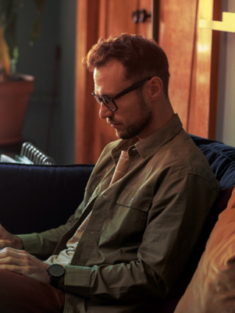 A person with glasses sits on a sofa, typing on a laptop. They are in a cozy room with warm lighting and a potted plant nearby.