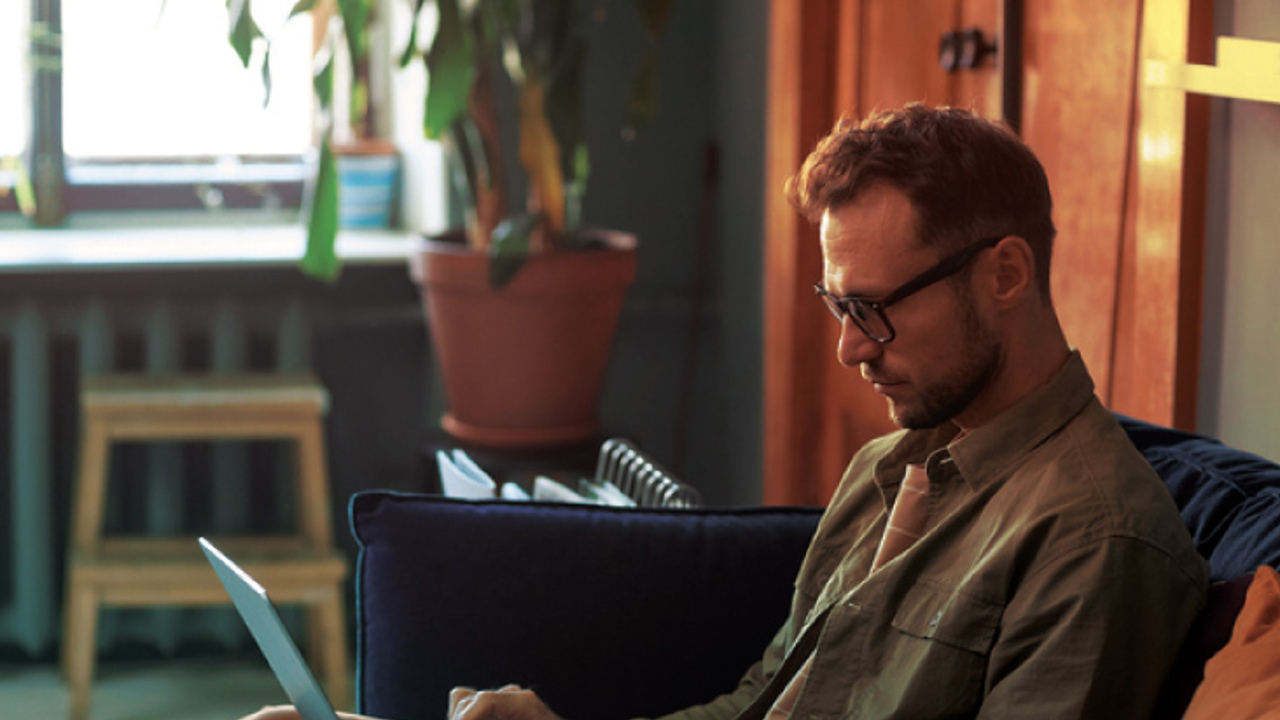 A person with glasses sits on a sofa, typing on a laptop. They are in a cozy room with warm lighting and a potted plant nearby.